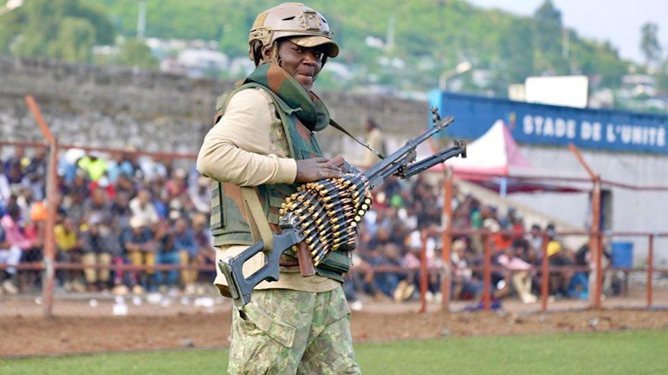 A M23 fighter holding a gun with lots of bullets wrapped around it. He is standing in a stadium with people seated behind him - Goma, DR Congo