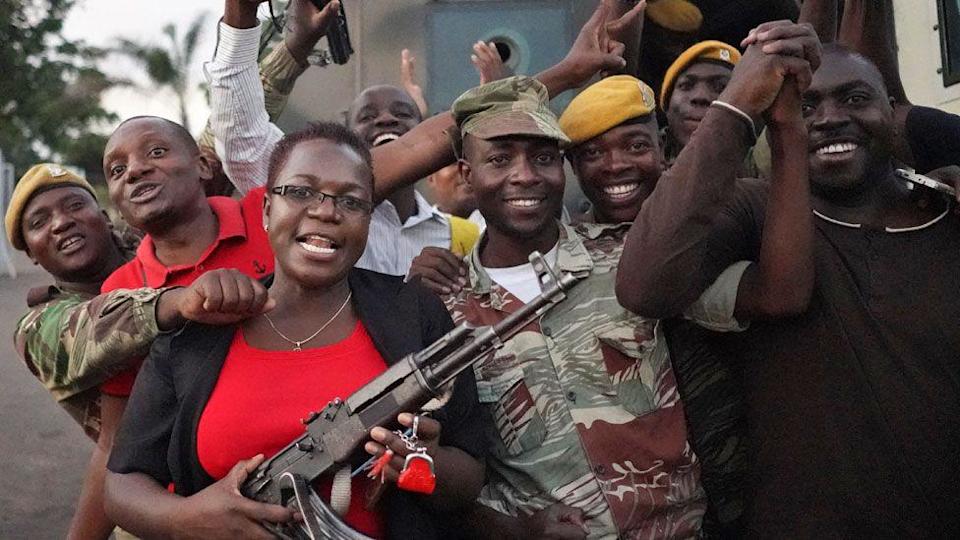 Zimbabweans celebrate with soldiers on the street including a woman in a red T-shirt and black cardigan holding a machine gun in 2017