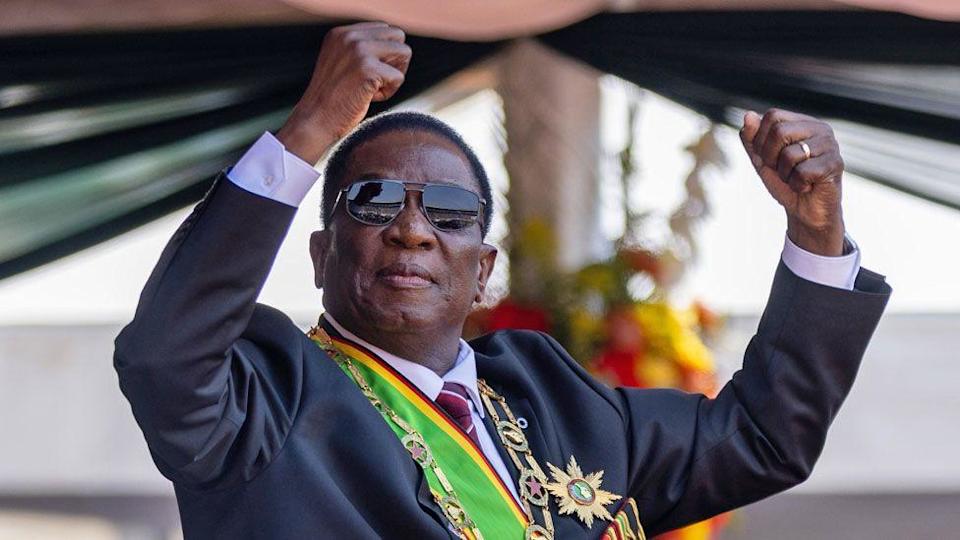 Zimbabwe&#39;s President Emmerson Mnangagwa in sunglasses and wearing a suit and sash in the colours of Zimbabwe and a chain and star of office holds up his fists.