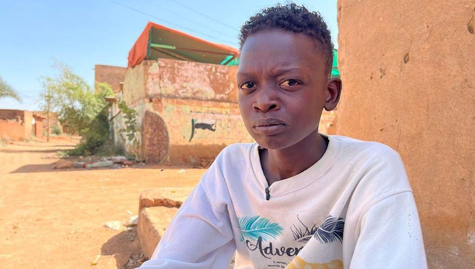 Muzami, the 18-year-old son of Intisar Adam Suleiman, looks directly into the camera lens with a pained expression. He is wearing a white sweat shirt and sitting outside an orange-plastered building on a street in the Haj Yusuf district of Sudan&#39;s capital Khartoum - March 2025