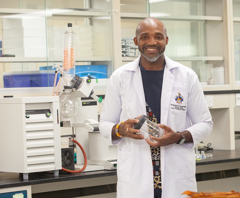 Brian Elcock smiles as he stands in his lab wearing a white lab coat