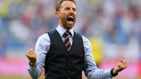 PA Media Gareth Southgate, a man in a pale blue suit and navy waistcoat, celebrates with his fists clenched and his mouth wide open on a football pitch
