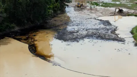 Area of light brown water with black, oily-looking substance along the bank and partially along a containment boom that runs across the water. Black mud and two diggers can be seen in the background.