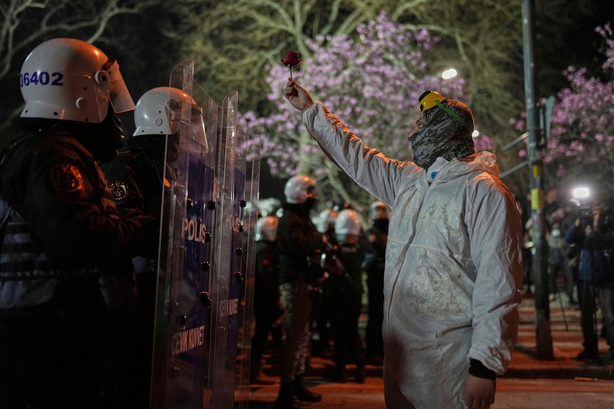 A person in a black hazmat suit holds up a flower in front of police in riot gear 