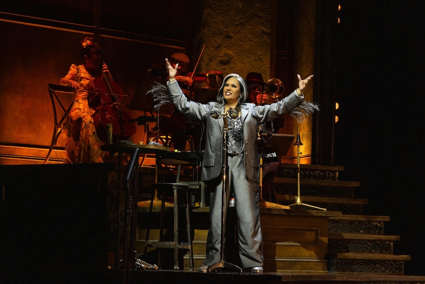 Christine Anu, in silver jacket and pants, holds hands up in the air victoriously, smiling, with people on stage behind her.