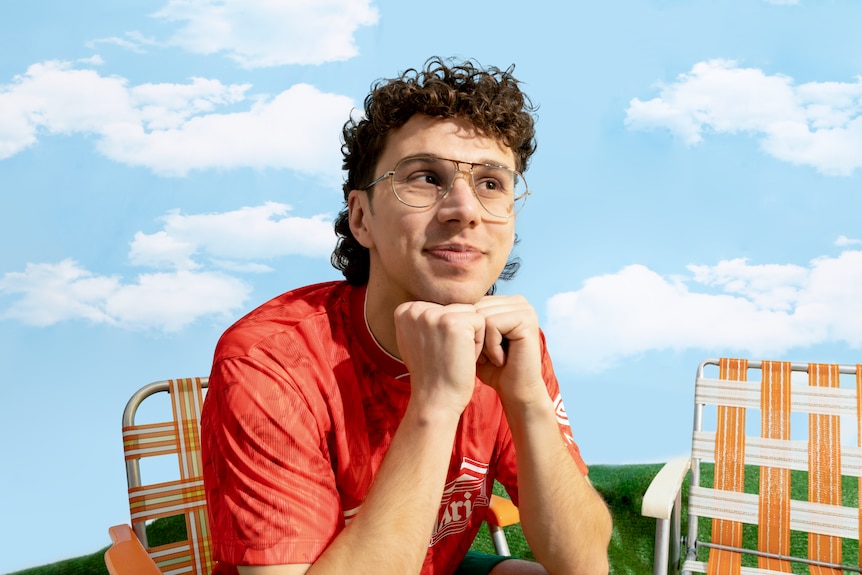 A man with curly short hair and glasses in a red shirt sits on a deck chair with his hands clasped under his chin