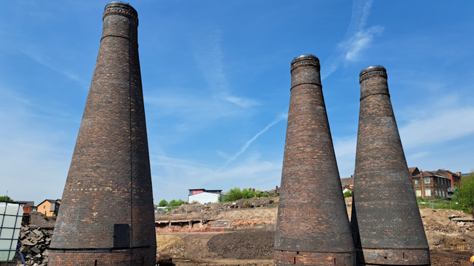 Three bottle kilns in a line on a building site with houses and trees in the background.