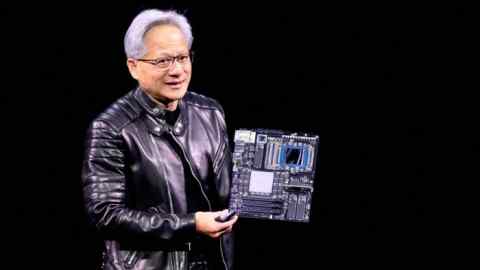 Jensen Huang holds up a circuit board while delivering a speech in a black leather jacket and standing against a dark background