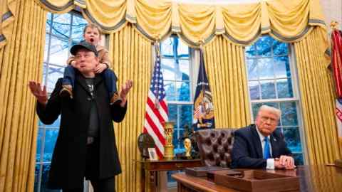 Elon Musk stands with his son X Æ A-Xii on his shoulders as Donald Trump sits at the desk, looking on