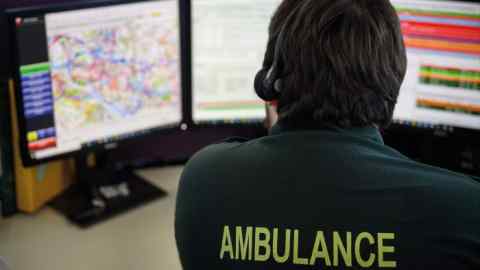 A person in an ambulance uniform sits at a workstation with three computer monitors displaying maps and emergency dispatch data. The person is wearing a headset