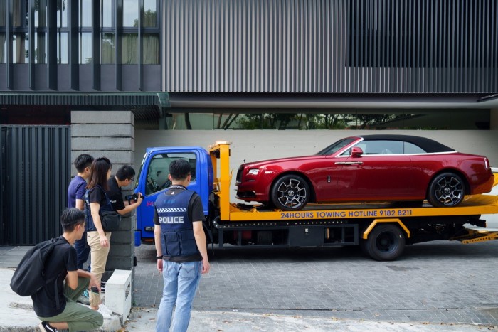 A red Rolls-Royce is loaded on to a flatbed tow truck in front of a modern building, with several people, including a police officer, observing and documenting the scene