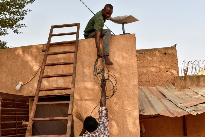 A man is on the roof holding the dish, next to a ladder. Another man on the ground is passing cabling up to him